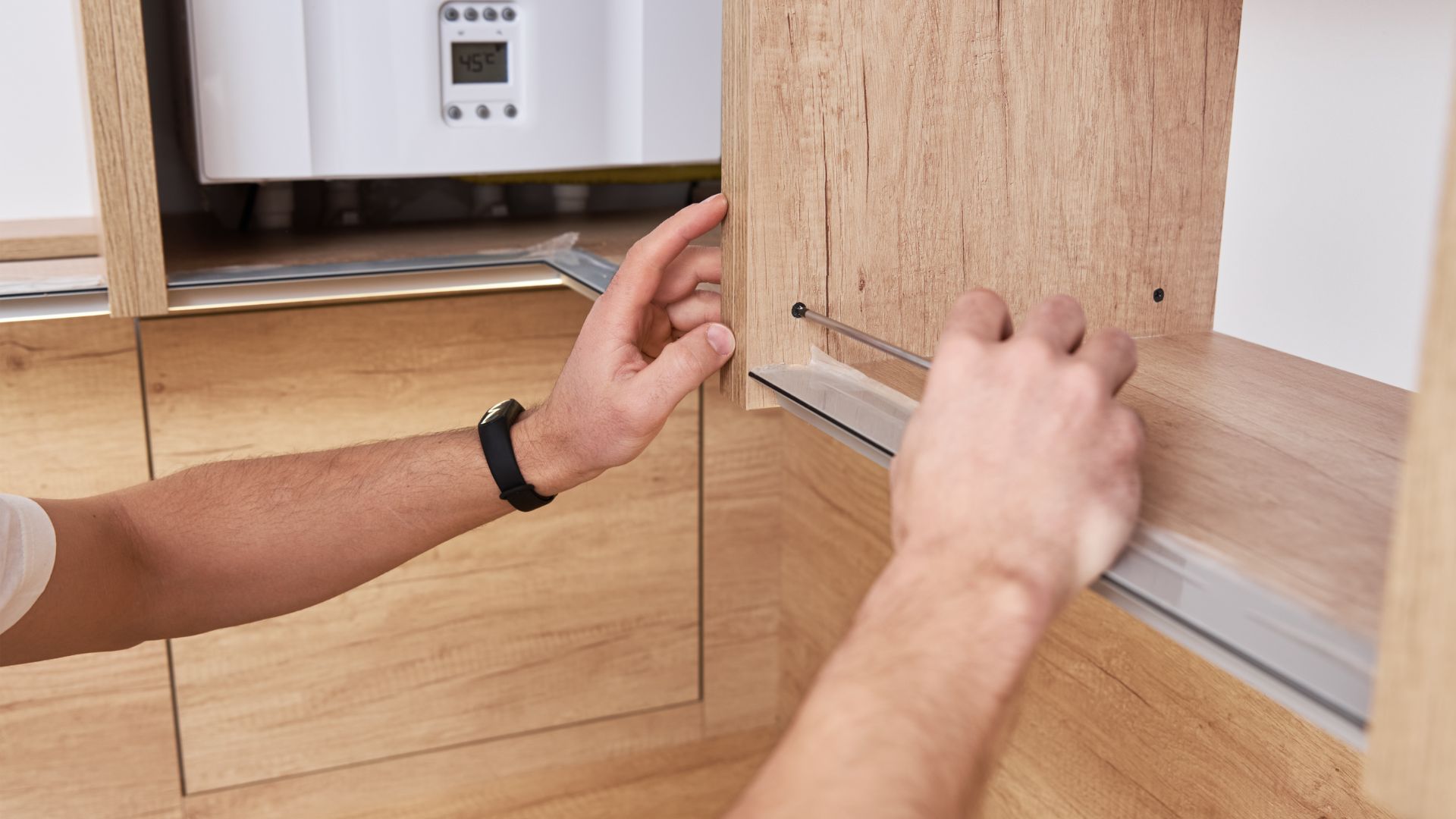 Hands installing or adjusting wooden cabinet with digital thermostat in background