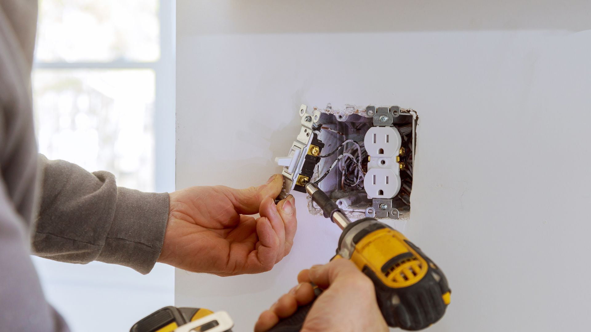 Electrician installing electrical outlet with screwdriver near window